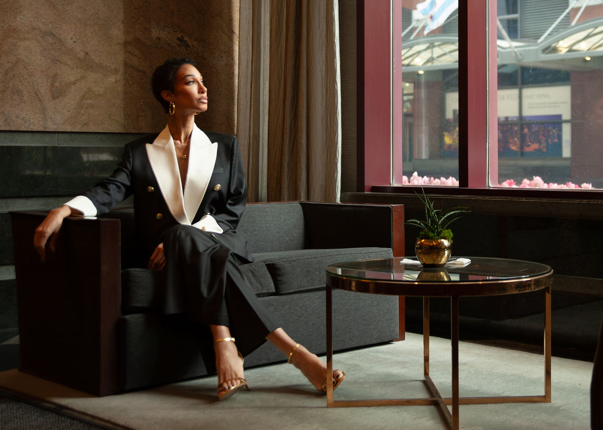 The Mayfair Blazer and Mayfair Pants by UNROOTD CHAPTRS, black wool with white silk lapels, worn seated in a hotel lounge by a window